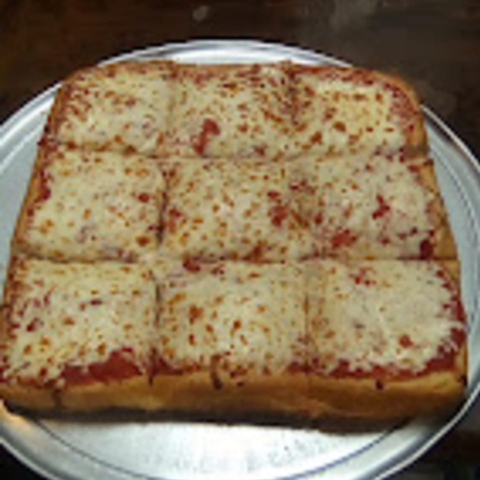 A rectangular Brooklyn Pizza cheese pizza, cut into nine squares, on a metal pizza pan.