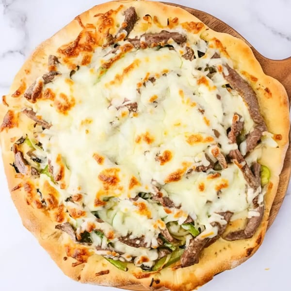 Cheesesteak pizza with melted mozzarella, sliced steak, and green peppers on a wooden peel.