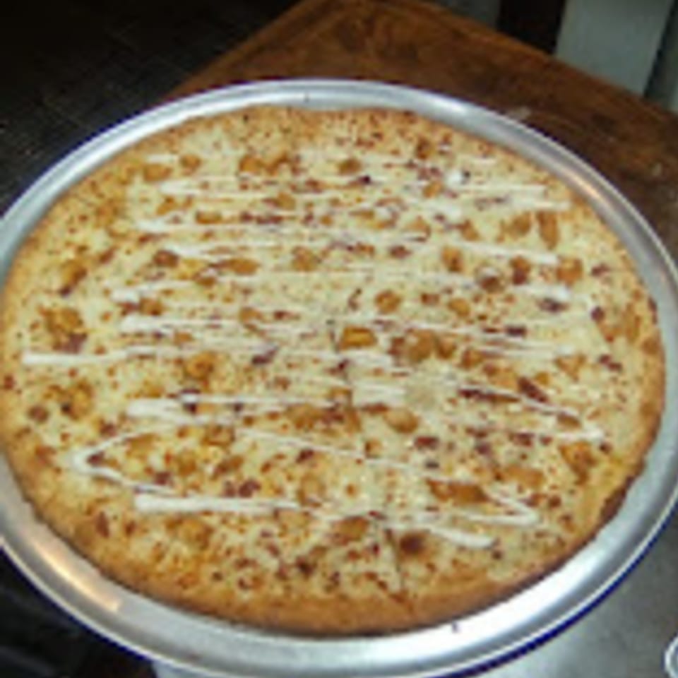 A whole Brooklyn Pizza chicken bacon ranch pizza, drizzled with white sauce, on a metal pan.
