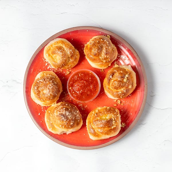 Six golden-brown garlic knots dusted with Parmesan cheese, surrounding a cup of marinara sauce on a red plate.