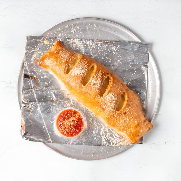 A golden-brown calzone with visible cheese and sauce, served on foil with a side of marinara.